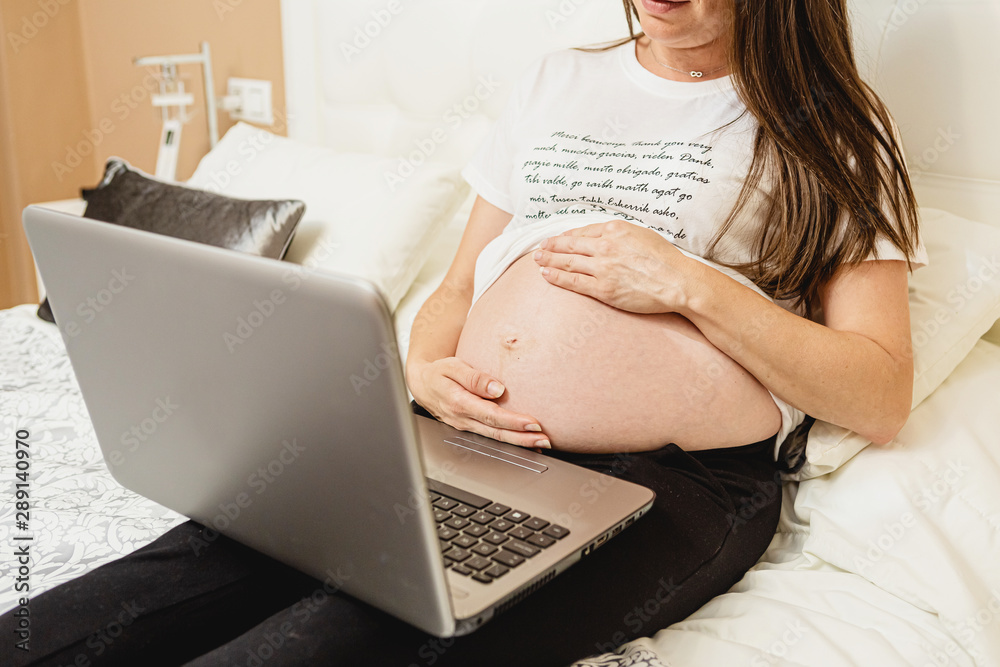 Naklejka premium Close up of a beautiful pregnant young woman showing her belly and working on her laptop at home