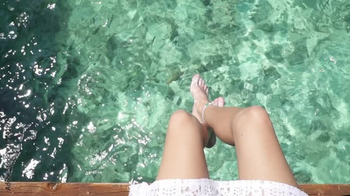 Young lady sitting on wooden jetty with crossed legs swinging above turquoise ocean. Maldives. Water bungalows. Travel motivation. Relaxing on resort. Super slow motion.
