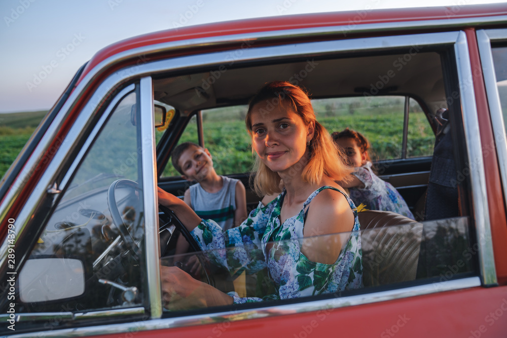 Close up photo of a beautiful young blonde mom in dress with flowers print driving a car with her teen children, enjoying traveling together