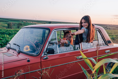 Beautiful young mom amd her daughter traveling together by the car, both in blue dresses, mom is driving and her teen daughter got out of the car window and pointing into the distance