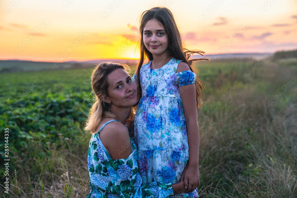Young blonde mom sat down near her daughter and hugging her, both wearing dresses with flowers print, standing among the mealow and looking at the camera, sunset on the background