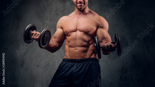 Happy smiling bodybuilder is doing exercises with barbels and showing his muscules on the dark background.