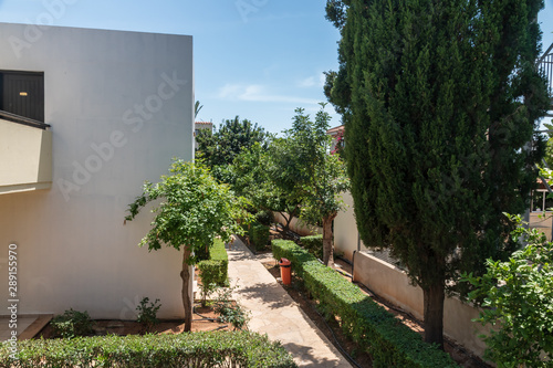 The walkway in the hotel with green bushes. Light building and entrance to the hotel room. Green trees with summer blue skies.