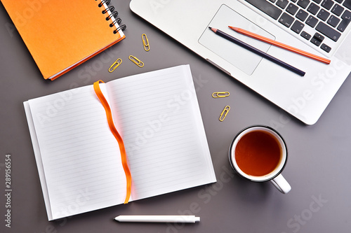 Top view of gray office desk with opened notebook or diary with blank page, cup of tea and laptop. Business concept. Copy space