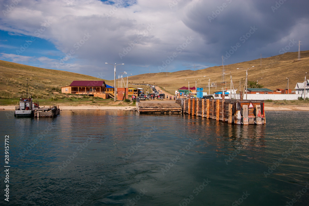 Ferry pier Olkhon island with waiting cars and people. On the shore are wooden houses, lampposts. Over the hills clouds.