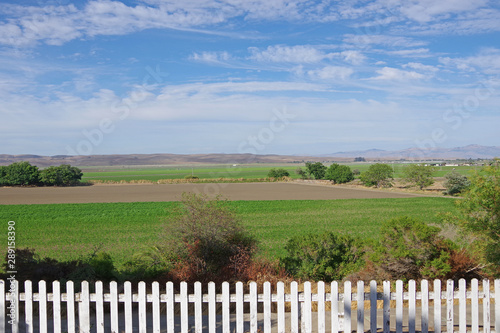 A panoramic view of the green California valley landscape along the St. Andreas fault with a white picket fence in front and blue sky