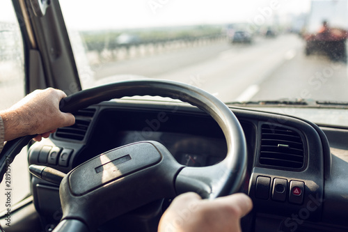 Male driver hands are holding steering wheel of truck during the movement in the road. Image with selective focus on the wheel, dashboard and blurred windshield