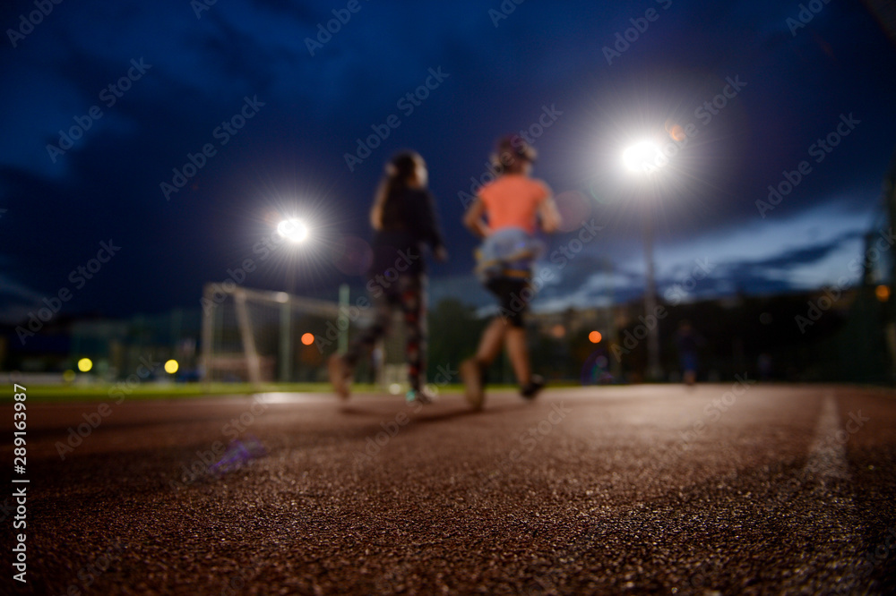 two young sport females running on outdoor stadium track at twilight under bright lights
