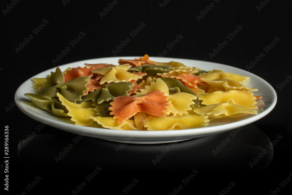 Lot of whole red, yellow and green uncooked farfalle on white ceramic plate isolated on black glass Lot of whole red, yellow and green uncooked farfalle on white ceramic plate isolated on black glass