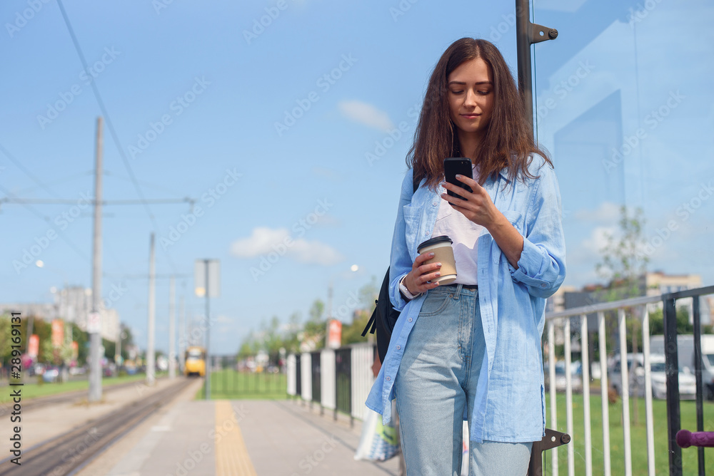 Pretty girl with cup of coffee waits for bus or tram on public transport station in the morning. Young woman with smart phone monitoring transport through the app.