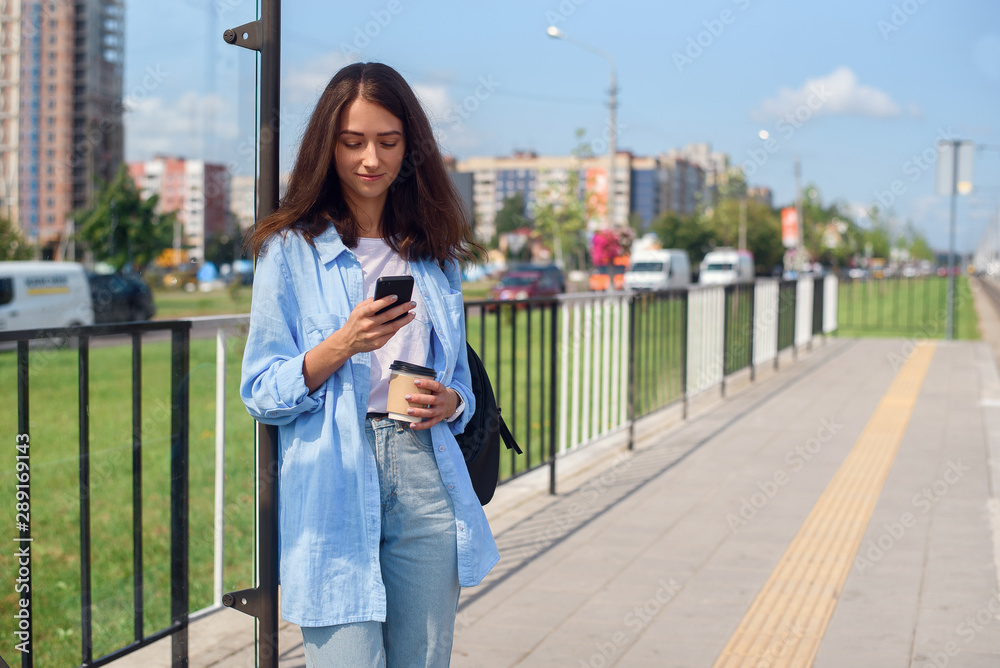 Fototapeta premium Pretty girl with cup of coffee waits for bus or tram on public transport station in the morning. Young woman with smart phone monitoring transport through the app.