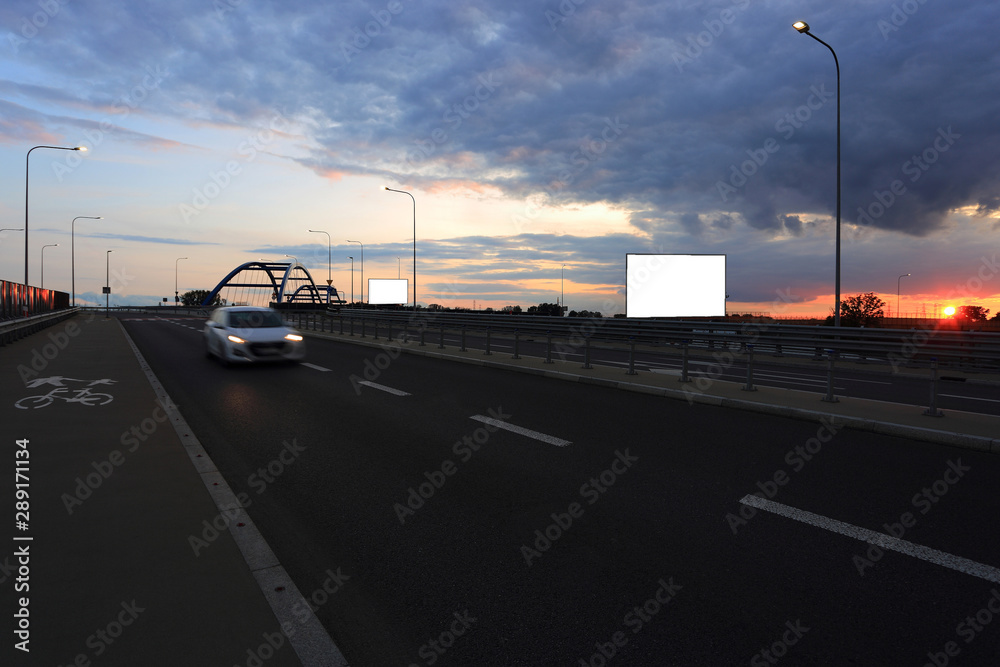 Samochód osbowy na drodze, zachód słońca, bilbord reklamowy, tekst, wiadukt, most.	