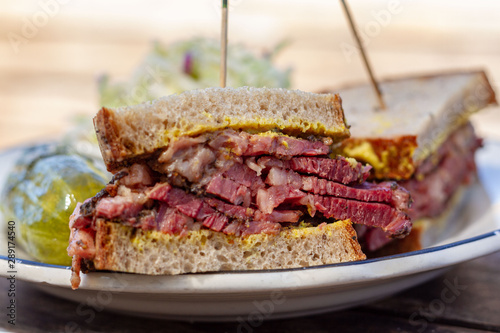Fotografie Pastrami sandwich sliced in half on rye bread with mustard on a white plate on a wood table as seen from above