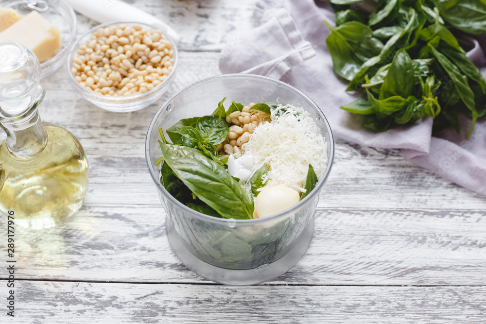 The process of making pesto sauce in a blender from cheese, pine nuts, olive oil, basil, garlic on the white wooden table. The process of making pesto sauce in a blender from cheese, pine nuts, olive oil, basil, garlic on the white wooden table.