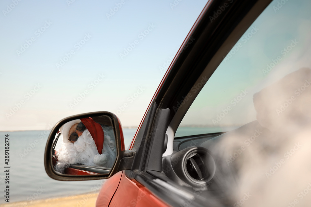 Authentic Santa Claus looking into side view mirror of car near sea