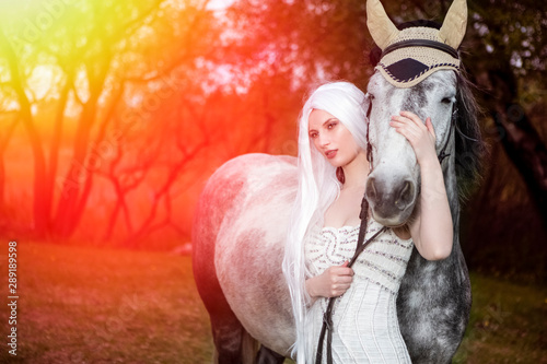 Beautiful Caucasian Blond Female with long Hair Standing with Grey Horse Against Bright Light on The Background