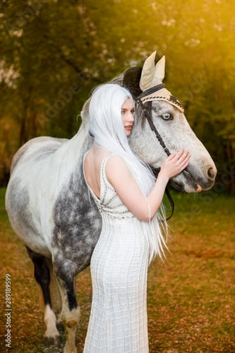 Sensual and Sexy Caucasian Blond Female with Long White Hair Standing with Horse Against Bright Light on The Background.