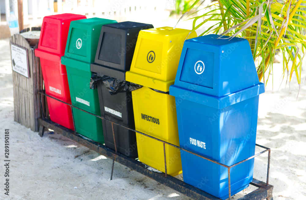 Garbage trash bins for waste segregation. Separate waste collection food  waste, infection, biodegradable, non biodegradable and recycle trash bin.  Multicolor trash bin on a sandy beach under coconut. Photos | Adobe Stock