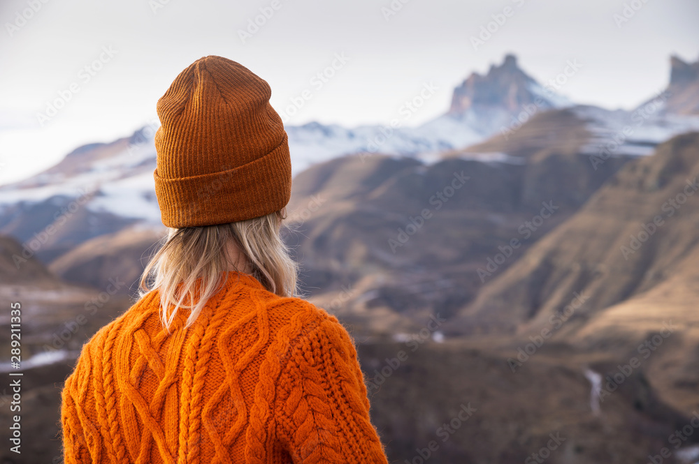 Portrait from the back of the girl traveler in an orange sweater and hat in the mountains against the background of a frozen mountain. Photo travel concept Portrait from the back of the girl traveler in an orange sweater and hat in the mountains against the background of a frozen mountain. Photo travel concept