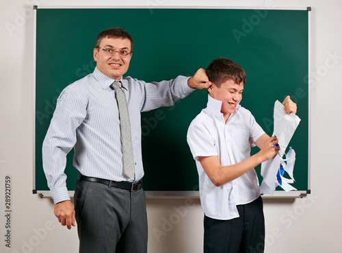 Portrait of a teacher catch the ear funny schoolboy with low discipline. Pupil very emotional, having fun and very happy, posing at blackboard background - back to school and education concept