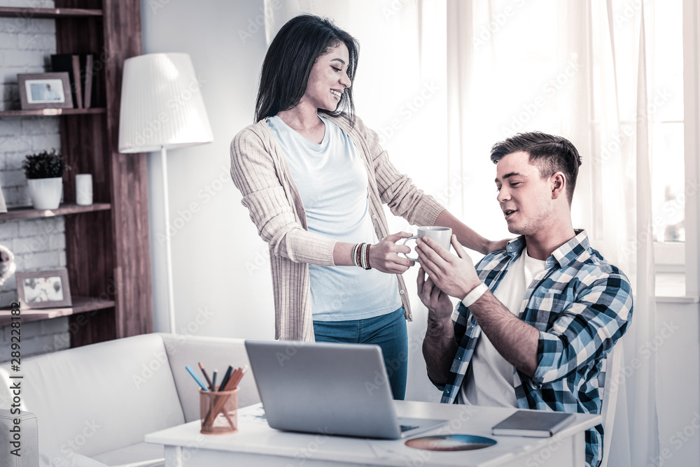 Obraz premium Caring young girlfriend giving cup of tea to her working boyfriend