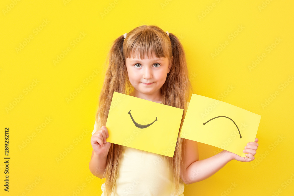 Little girl with drawn smiling and upset mouths on sheets of paper against color background Little girl with drawn smiling and upset mouths on sheets of paper against color background