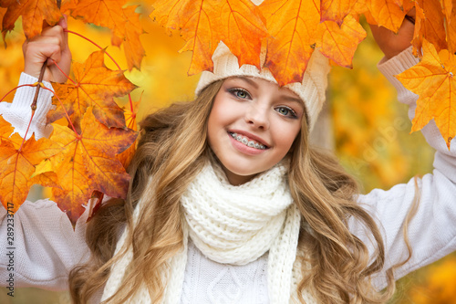 Portrait of beautiful young woman walking outdoors in autumn. Girl with autumn yellow leaf in knitted hat and sweater.  Portrait of a girl with orthodontic appliance.