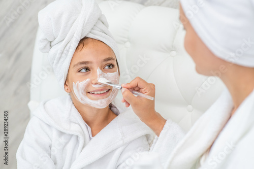 Mom puts white cream on her  smiling daughter's face at home