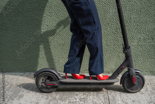 Young woman in formal wear on red hight heels is standing on electrical scooter. Close-up of female legs. A business woman in a trouser suit and red shoes moves around the city on an electric scooter.