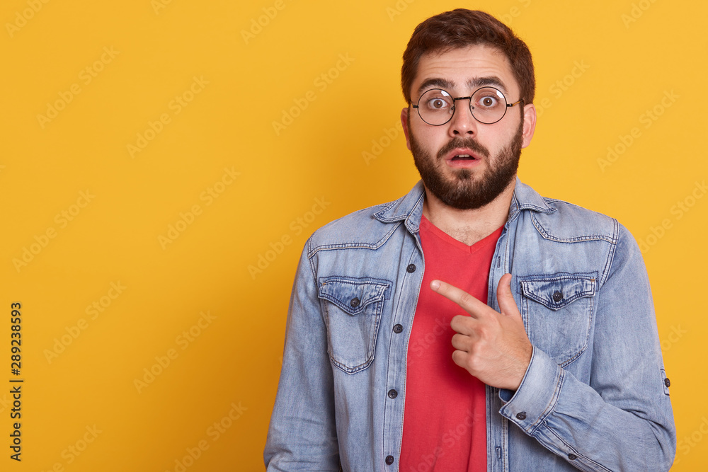 Image of shocked young man pointing finger aside at blank copy space, astonished surprised guy dressescasual clothing, looking directly at camera, standing isolated over yellow studio background. Image of shocked young man pointing finger aside at blank copy space, astonished surprised guy dressescasual clothing, looking directly at camera, standing isolated over yellow studio background.