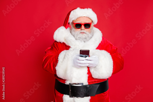 Portrait of minded santa claus with eyewear eyeglasses using cell phone device reading news wearing costume isolated over red background