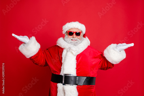 Portrait of handsome cool funny santa claus with eyeglasses holding newyear ads promo wearing black belt cap isolated over red background