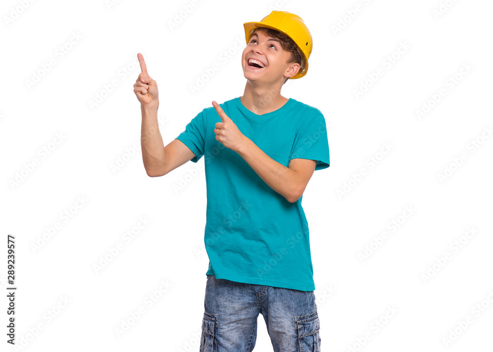 Portrait of teen boy wearing yellow hard hat pointing fingers away at copyspace, isolated on white background. Cute caucasian young teenager in helmet pointing fingers at something and looking up.