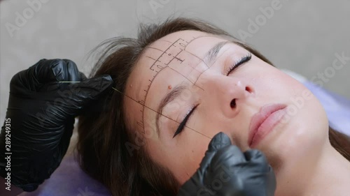 A face of a young lady and hands of a microblading master in black gloves. A specialist is marking the highest top of an eyebrow with a thread, a client is lying with closed eyes. Close up, portrait. photo