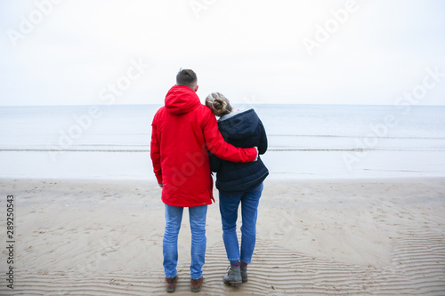 Fototapeta Naklejka Na Ścianę i Meble -  Two hipsters standing on the cold beach. Couple hugging and holding hands. Love story near the ocean. Winter season on the sea. Stylish boots on the sand. Man in red jacket.