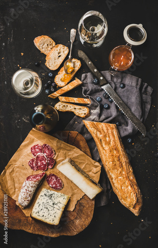 Wine and snack set. Flat-lay of sliced baguette, salami, cheese variety, apricot jam, berries and white wine over rustic black background, top view