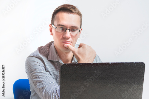 A young businessman in glasses is sitting at a table in the office with a laptop. Looks very thoughtfully at the screen.