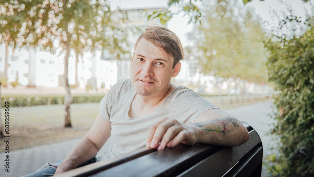 Fototapeta premium A young handsome man in casual clothes sits on a bench in a small alley.