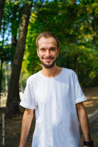 A portrait of a handsome man with beard against the background of green trees in the park.