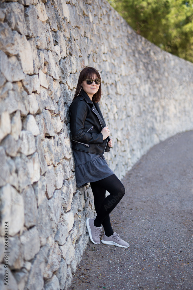 full length portrait of young woman in leather jacket and sunglasses over stone wall full length portrait of young woman in leather jacket and sunglasses over stone wall