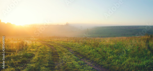 Sunny summer landscape with country road and green hills covered by morning fog