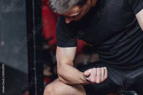 A young sportsman is sitting in a trunk hall. During this dress, wear a black T-shirt. Looking at your biceps hands. Serious.