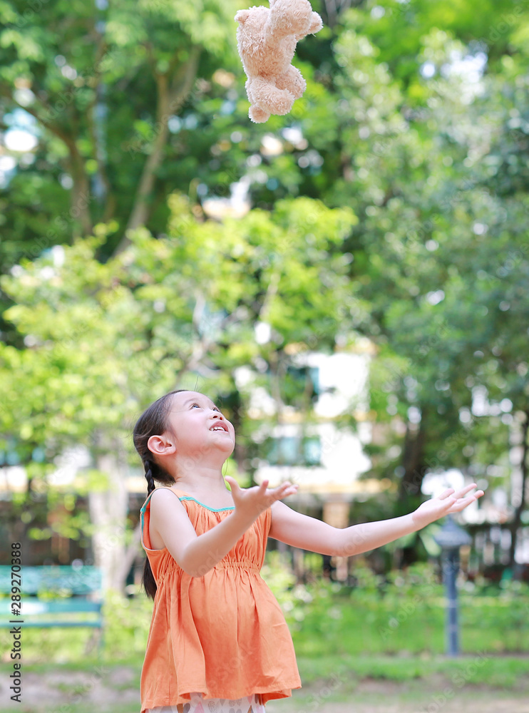 Portrait of happy little Asian child in green garden with throwing up teddy bear doll floating on air. Smiling kid girl playing in summer park. Portrait of happy little Asian child in green garden with throwing up teddy bear doll floating on air. Smiling kid girl playing in summer park.
