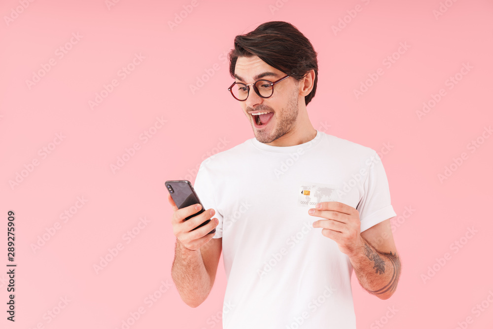 Image of handsome excited man wearing eyeglasses holding credit card while using cellphone Image of handsome excited man wearing eyeglasses holding credit card while using cellphone