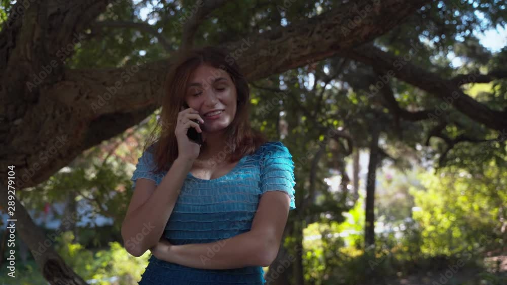 The girl in a blue dress happily speaks on the phone, smiles. Talking on the phone in the park. 4k The girl in a blue dress happily speaks on the phone, smiles. Talking on the phone in the park. 4k