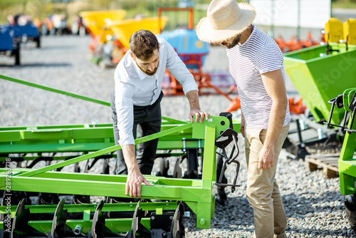 Young agronomist with salesman or manager at the open ground of the shop with agricultural machinery, buying a new plow for tillage photo