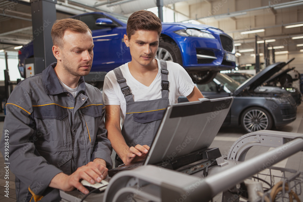 Two car service workers using computer, repairing broken automobile. Mature mechanic and his assistant doing computer diagnostics for a broken automobile