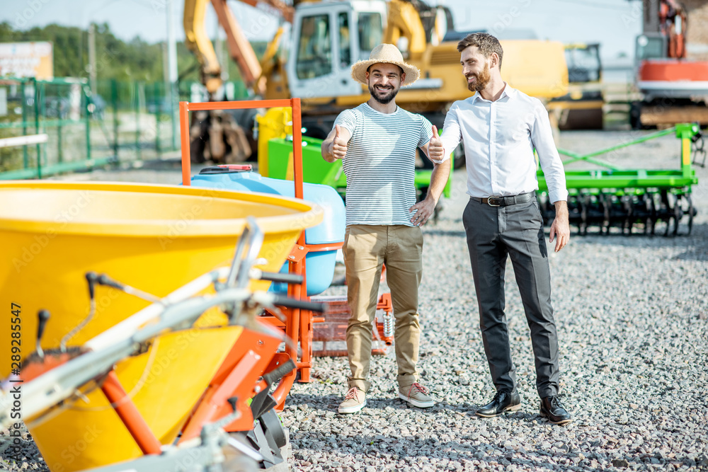 Young agronomist with salesman at the open ground of the shop with agricultural machinery, buying a new planter for farming