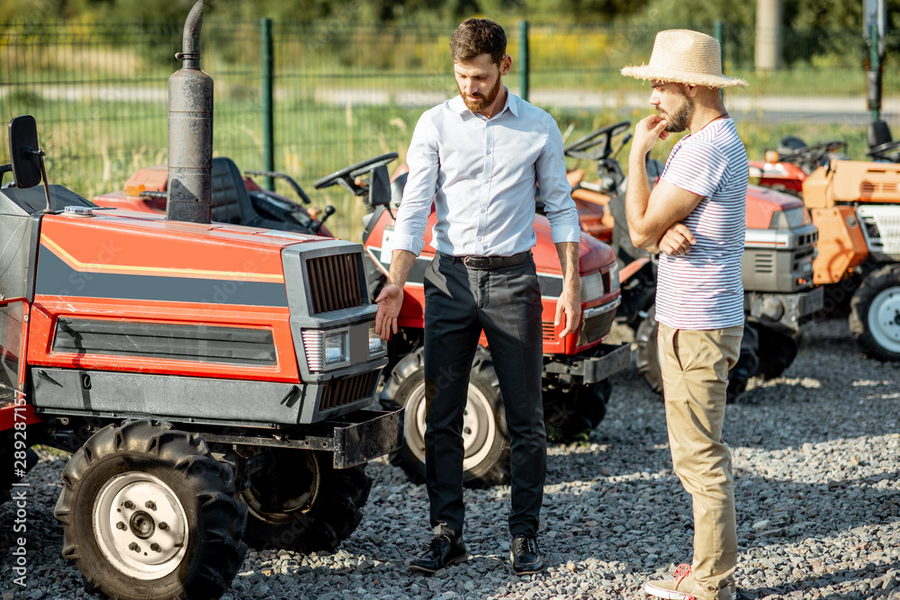 Obraz premium Young agronomist with elegant salesman choosing a tractor for farming on the open ground of agricultural shop