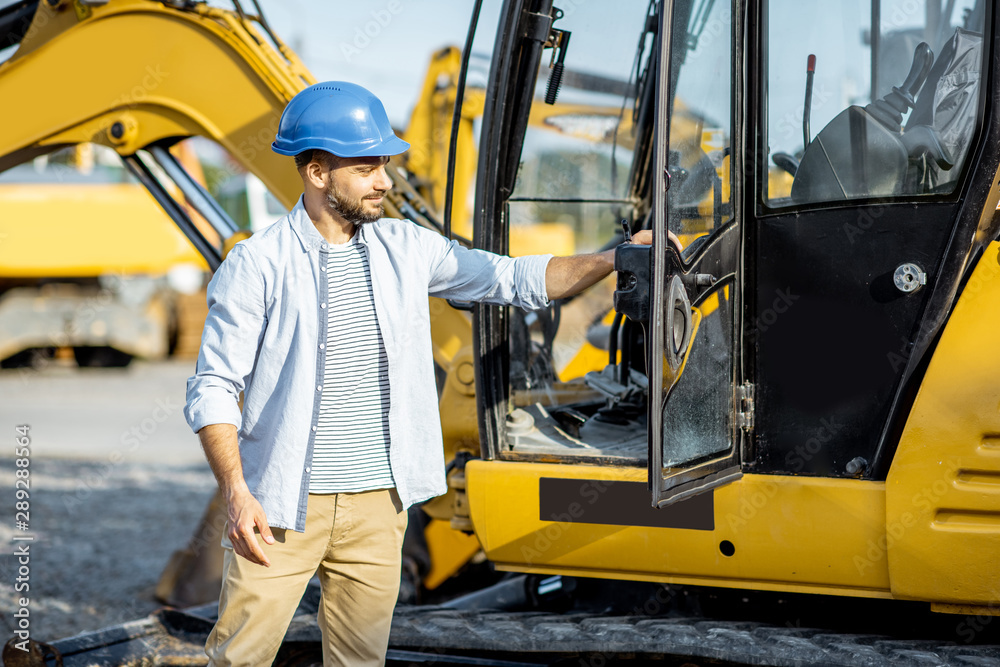Portrait of a handsome builder standing near the escavator on the open ground of the shop with heavy machinery Portrait of a handsome builder standing near the escavator on the open ground of the shop with heavy machinery